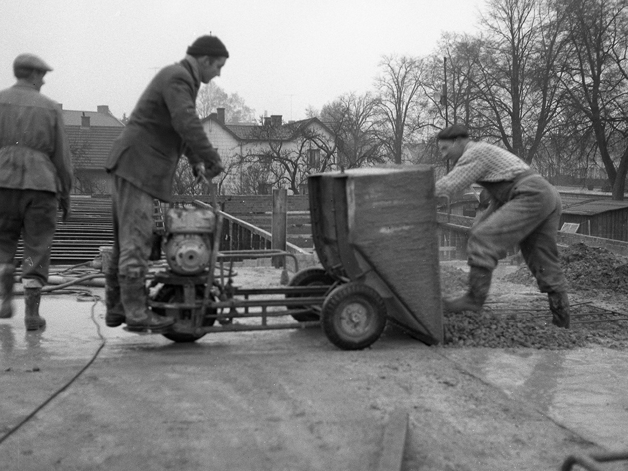 Two men in 1950s style work clothes and woolly hats are working with a large machine on a construction site.