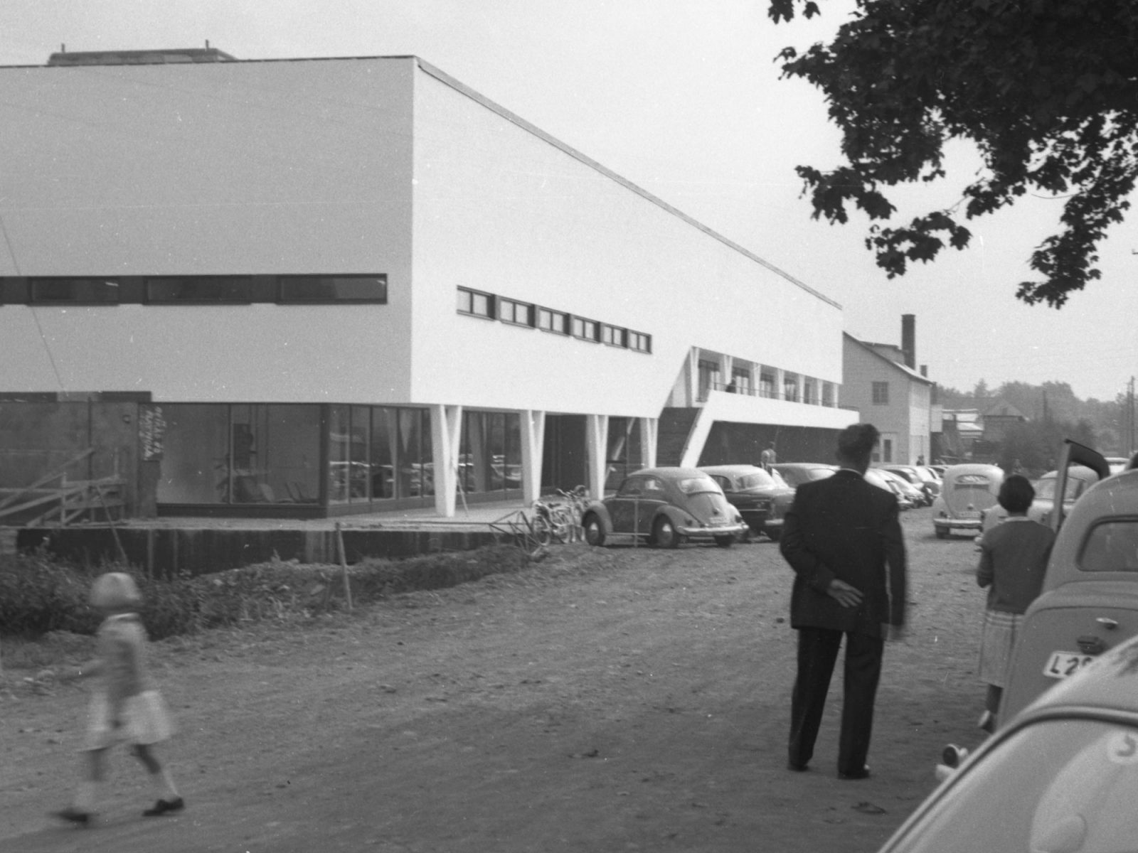 Family in 1950s style clothes step out of their car in front of large white modernist building, the first IKEA store.