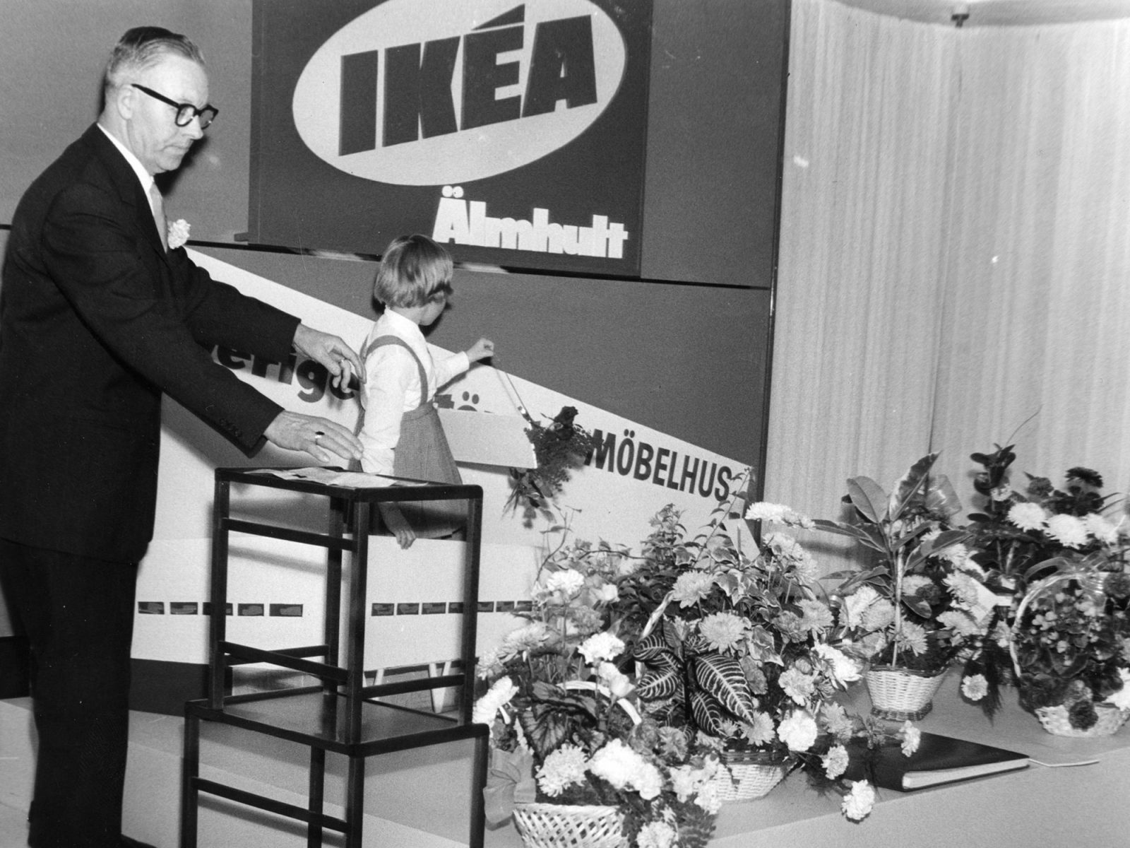 Un hombre mayor con traje oscuro, Eric Anderek, y una niña pequeña ante el cartel de IKEA; ramos de flores a sus pies.