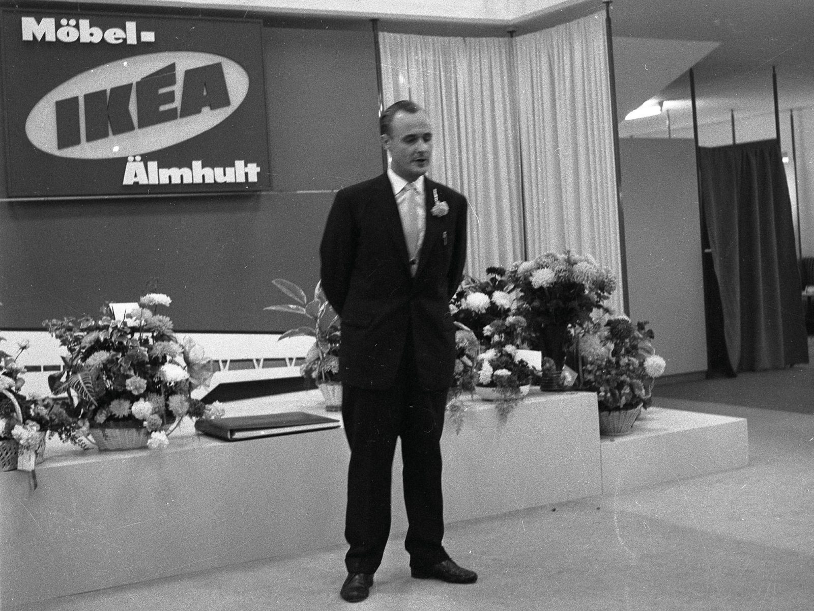 A young Ingvar Kamprad gives a speech wearing a dark suit in front of a large IKEA sign and lots of flowers.