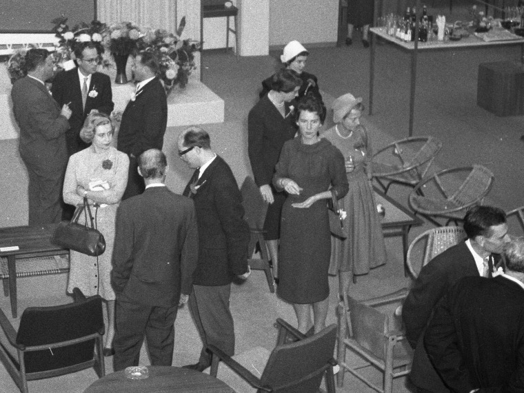 Des personnes discutent au milieu de meubles exposés. Les femmes en robe ou tailleur des années 1950, les hommes en costume.