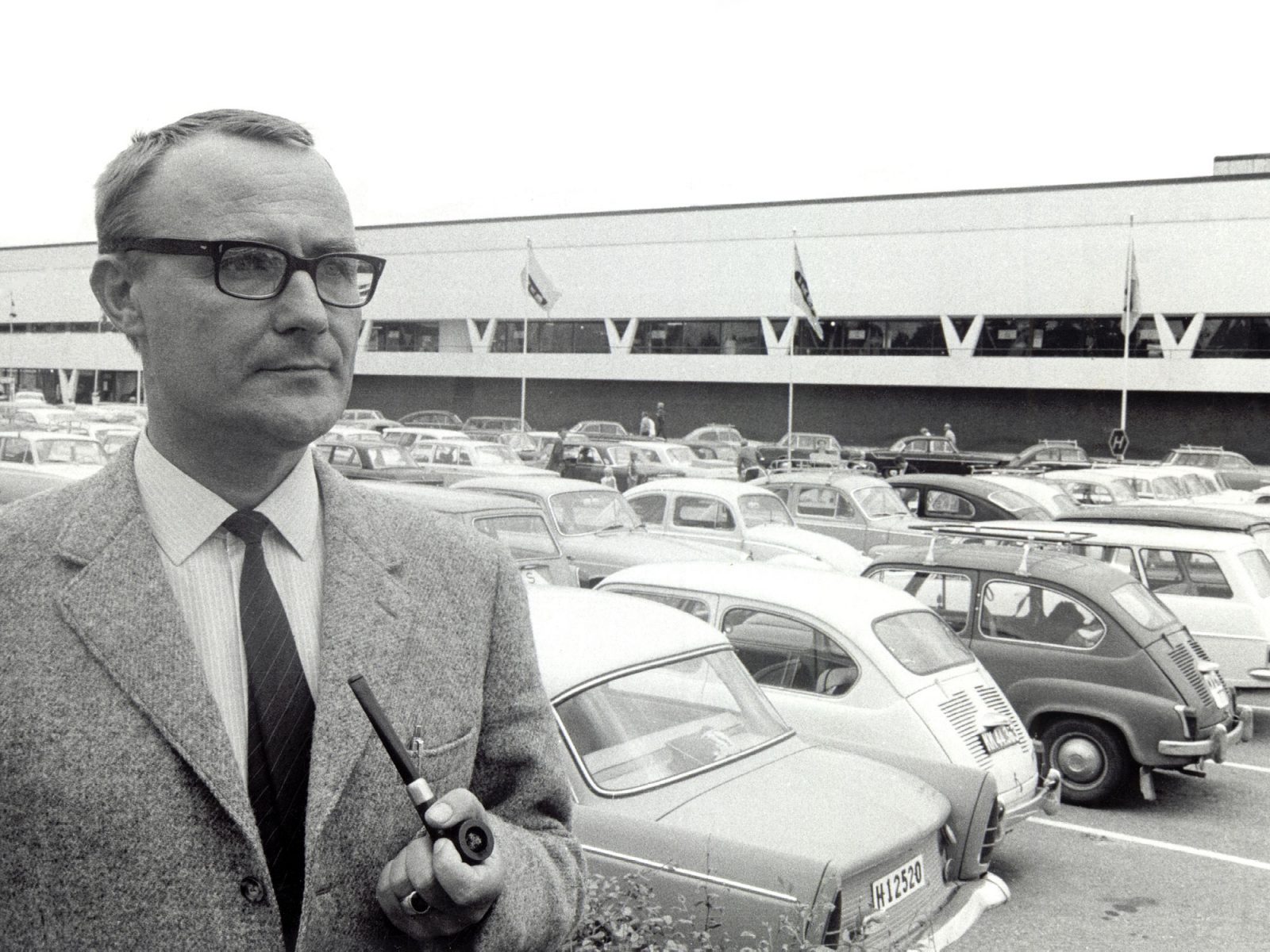Ingvar Kamprad en veste de tweed devant un bâtiment blanc, le premier magasin IKEA, parking avec voitures des années 1950.