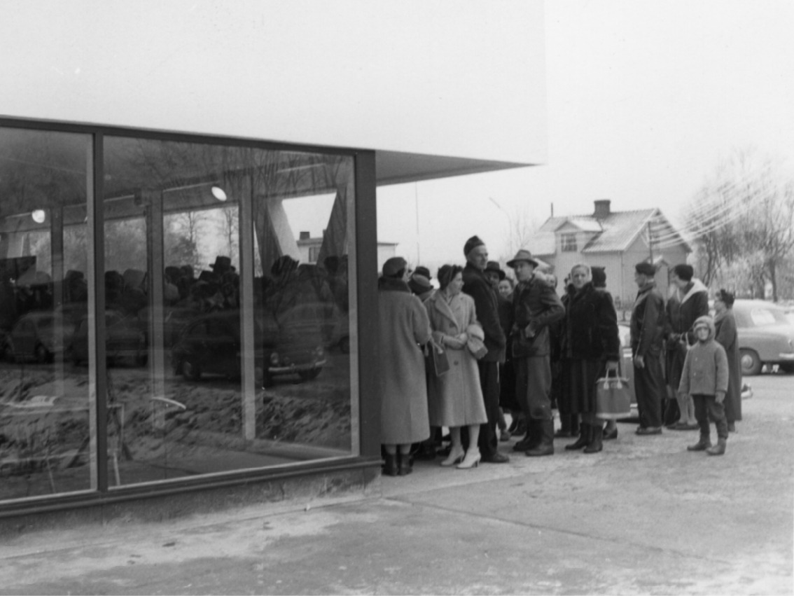 Menschen in 50er-Jahre-Kleidung stehen vor einem flachen Gebäude Schlange. IKEA in Älmhult, Schweden.