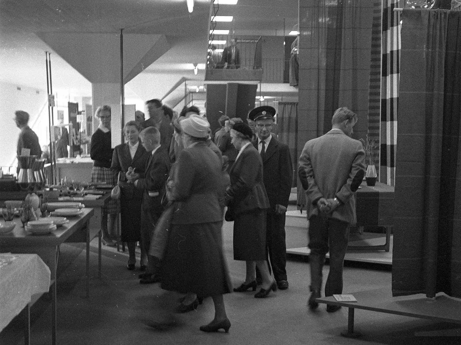 Des personnes discutent et regardent des meubles exposés. Femmes en robe ou tailleur des années 1950, hommes en costume.