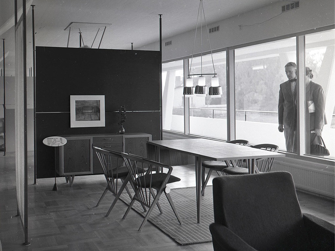 Jeune couple, style années 1950, regarde une vitrine exposant une pièce avec table, chaises, lampe et fauteuil.