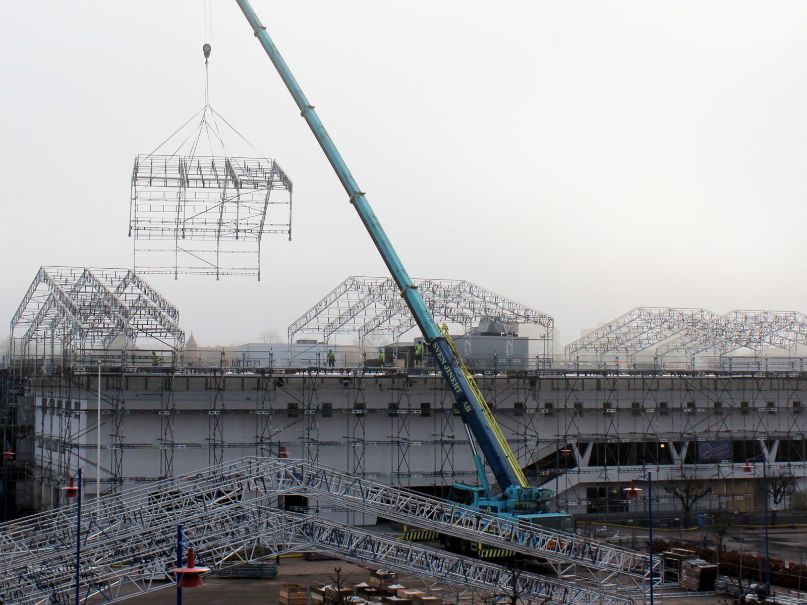 Översiktsbild visar en enorm byggarbetsplats, en stor kran lyfter upp byggställningar på ett tak.
