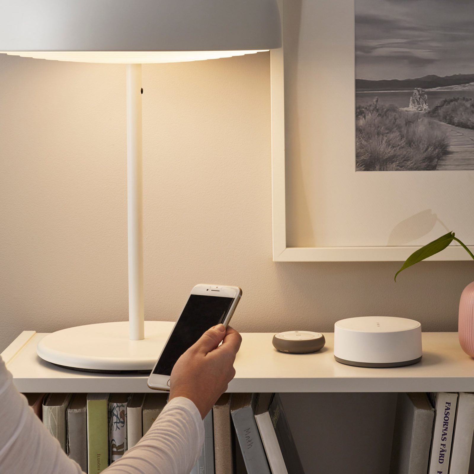 En person håller en smart phone framför en bänk med vit lampa och en liten rund vit wifi-gateway, TRÅDFRI.