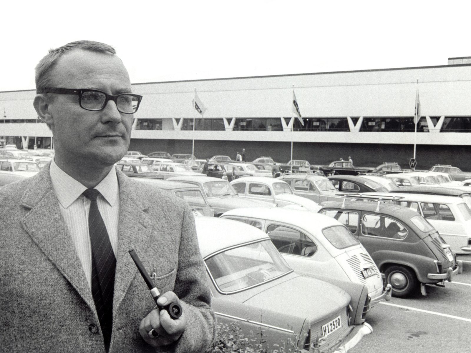 Ingvar Kamprad in tweed jacket stands in front of white building, the first IKEA store, and car park with 1950s cars.