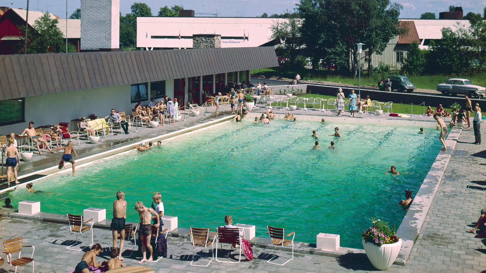Large pool full of bathing children next to sunbeds and motel building, Motell IKEA. 1950s cars in background.