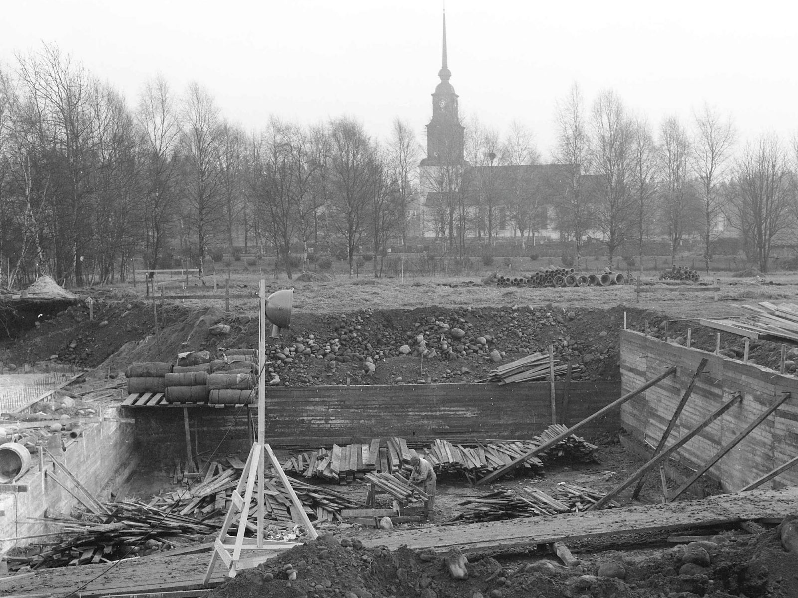 Byggarbetsplats, stor utgrävning för bassäng, en kyrka syns i bakgrunden.