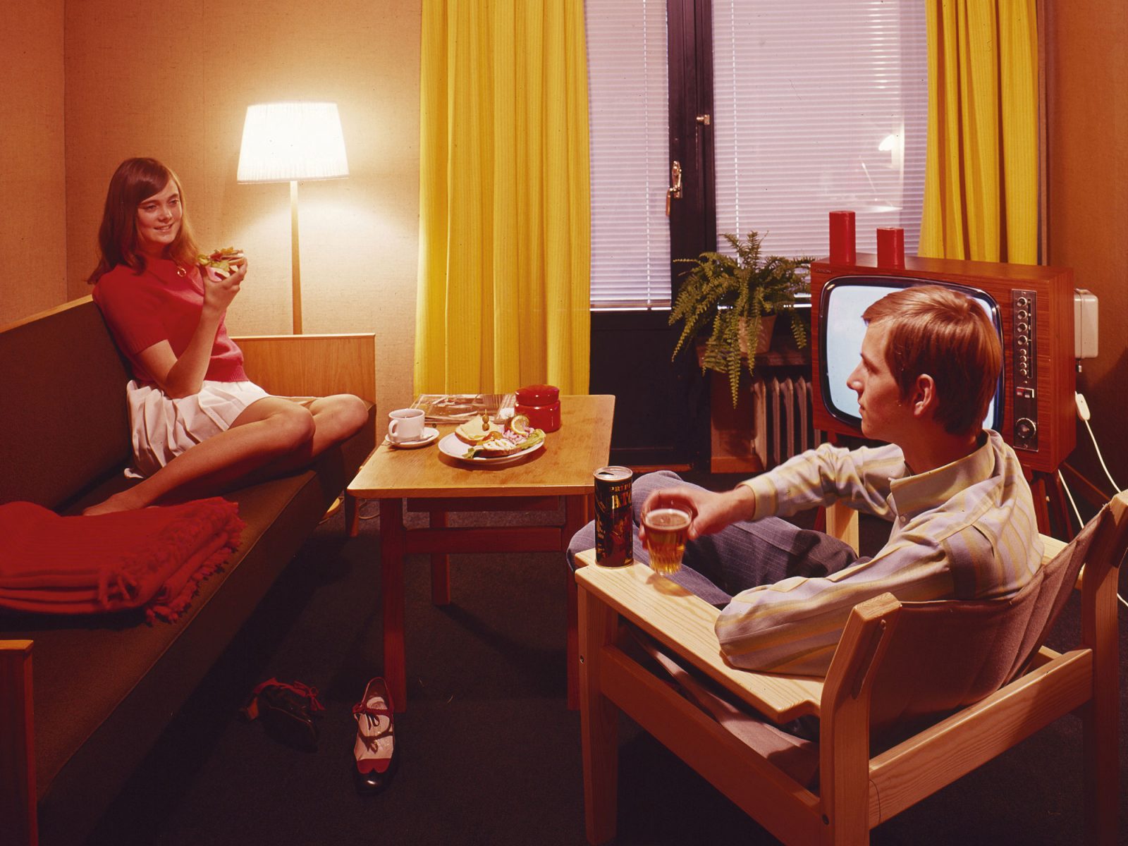 1960s style motel room interior, low oak sofa and yellow curtains, a couple drinking beer, coffee and eating snacks.