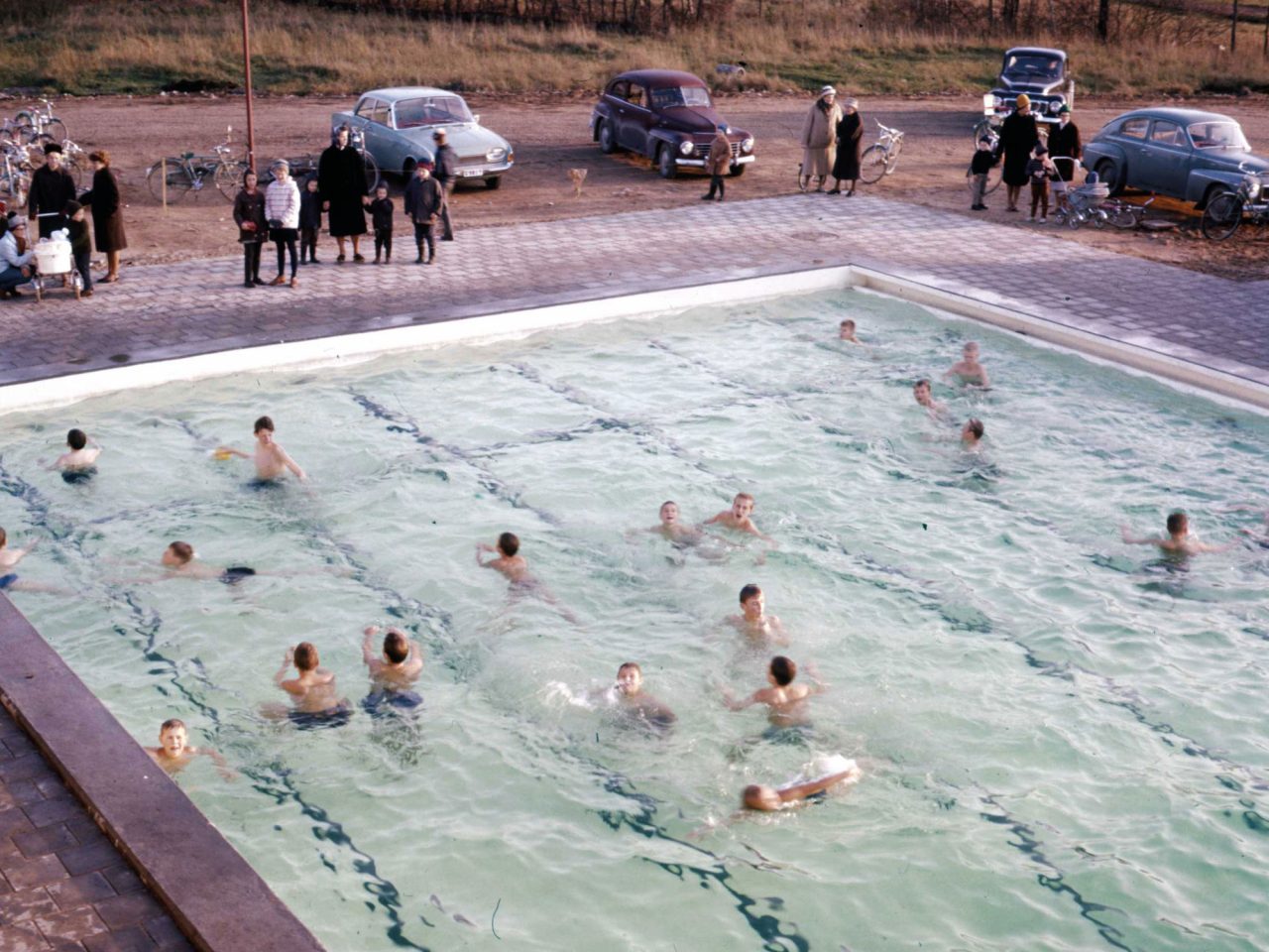 Large pool full of bathing children, people in warm clothing stand on the edge, 1950s cars seen in background.