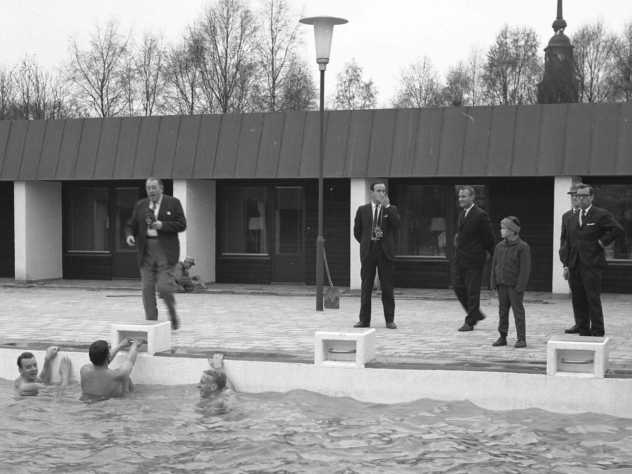 Men in suits, two with cameras, stand on the edge of a pool where children swim.