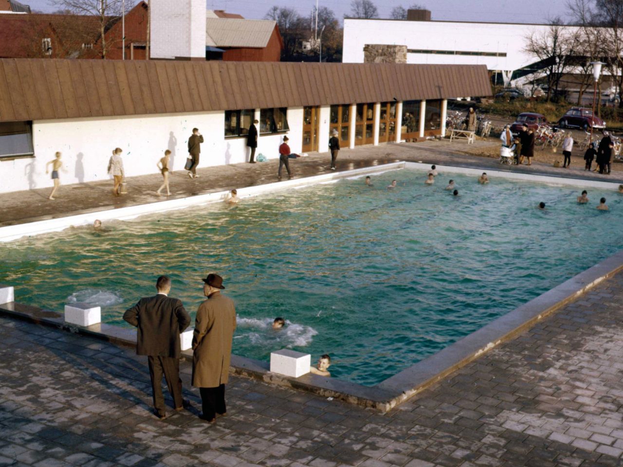 Image from above of swimming pool in autumn weather, two warmly dressed men in the foreground looking at children bathing.