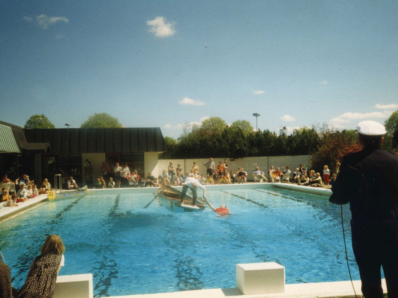 Publik runt badbassäng där man står i en liten roddbåt och visar livräddning av en person som ligger i bassängen.