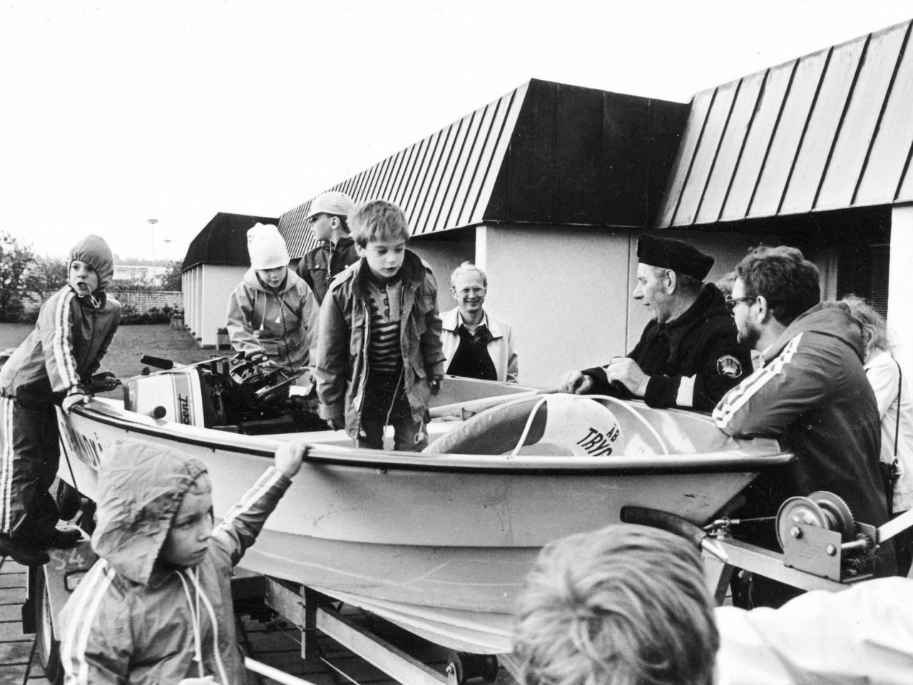 Children play in a boat set up on land, a policeman stands next to the boat talking to the children.