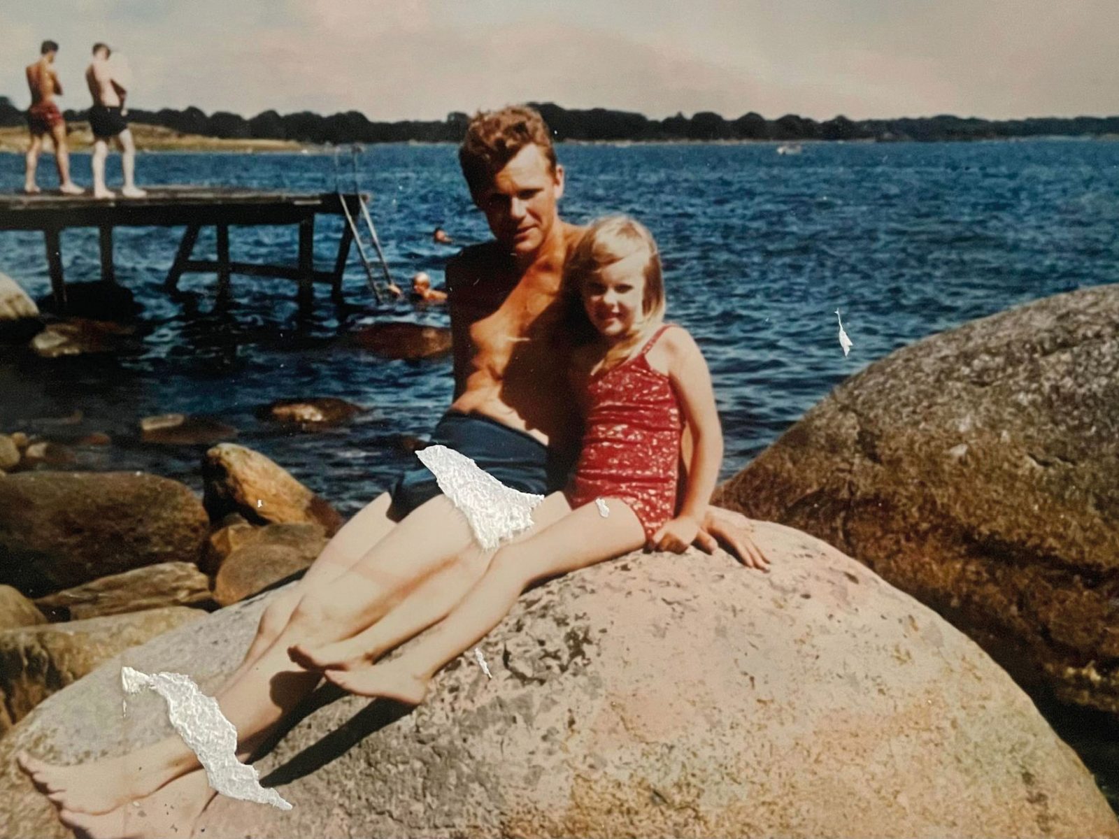Liten flicka i röd baddräkt, Ingela Johansson, sitter med man i badbyxor, hennes pappa, på stor sten vid badsjö.