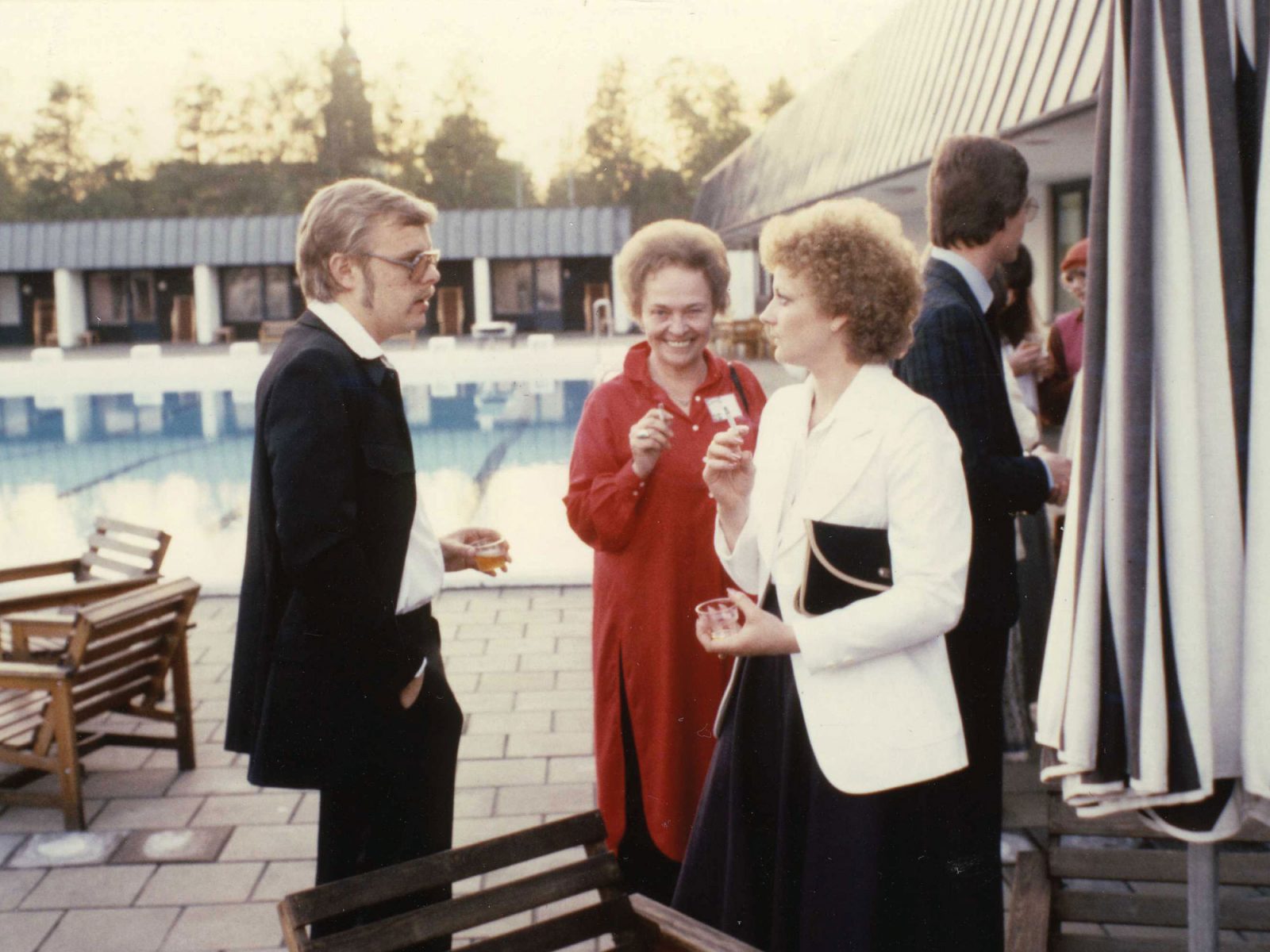 Tre personer minglar vid bassäng i 1970-talskläder, Jörgen Svensson, IB Bayley och Christina Folkinger.