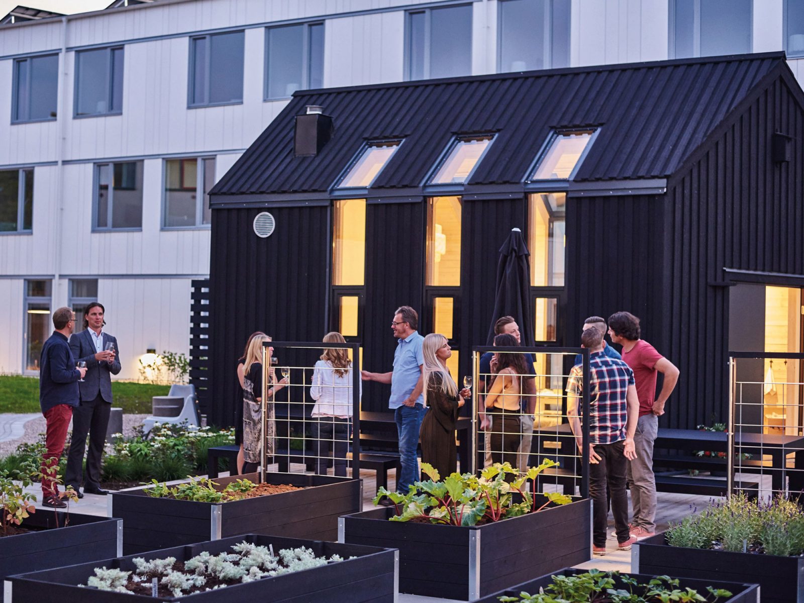 Casually dressed people mingling by a small black house, in the foreground you can see pallets with plants.