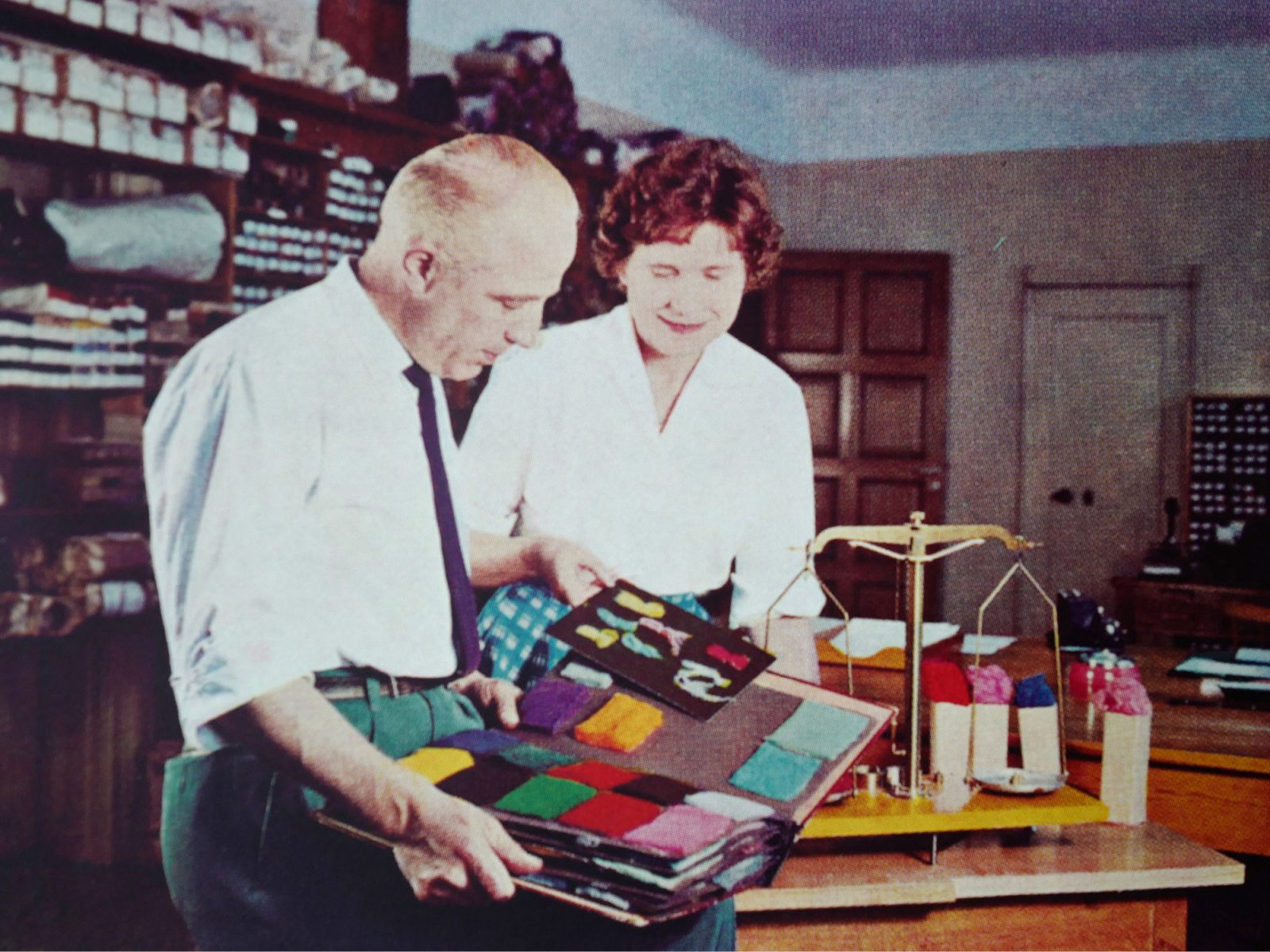 Kvinna och man, båda iklädda vita skjortor, tittar på färgglada tygprover i ett utställningsrum för textilier.