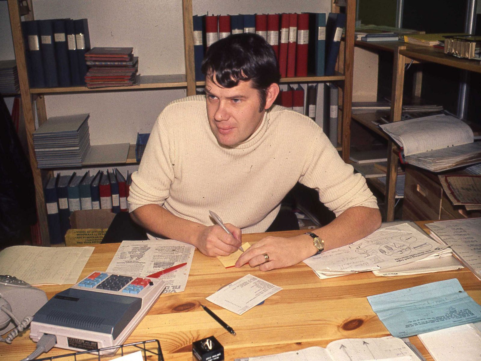 Lars Göran Peterson, ung man med polisonger i 1970-talsstil och vit stickad polotröja arbetar vid skrivbord.