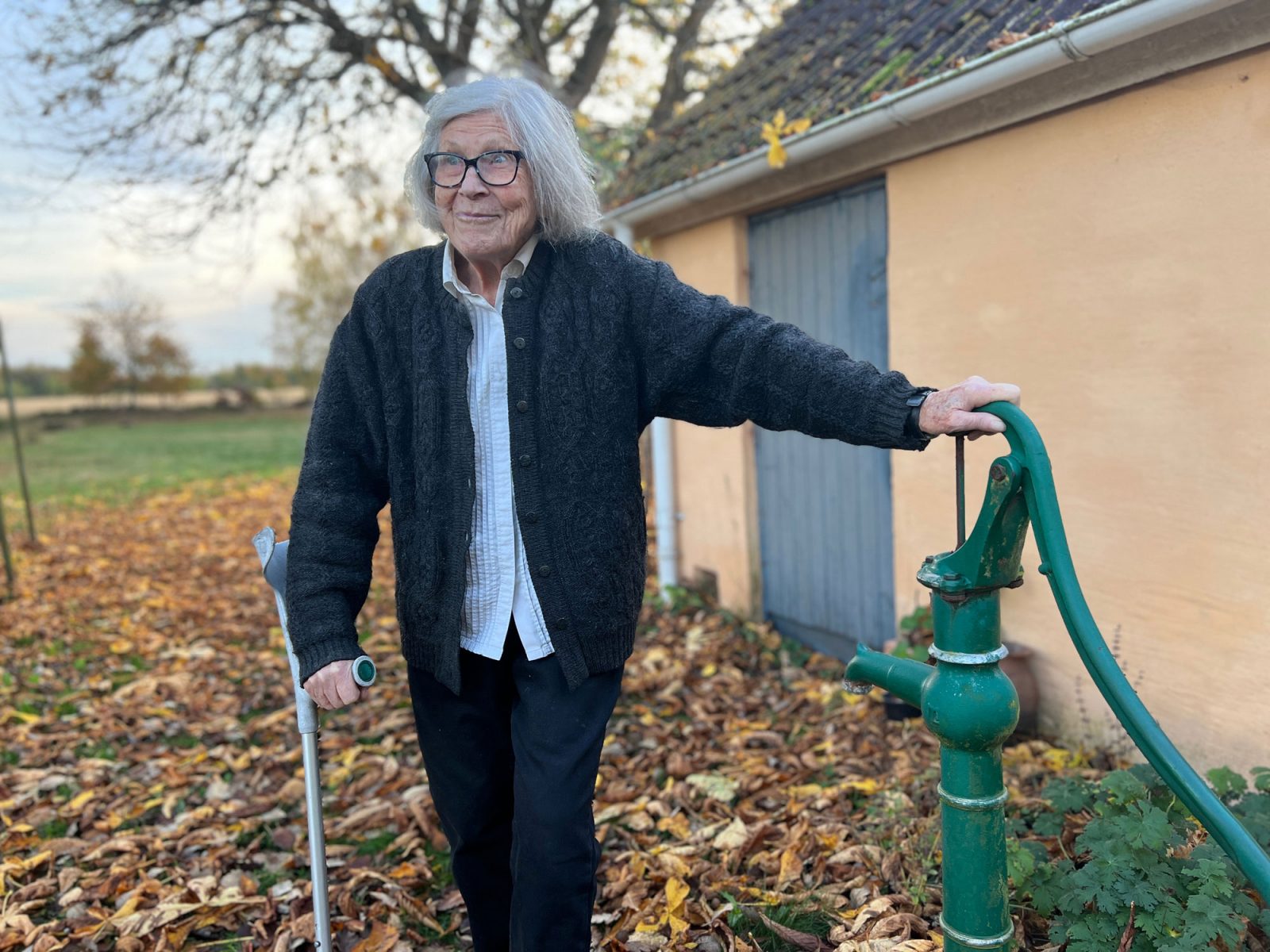 Elderly grey-haired woman standing in front of yellow house, next to water pump.