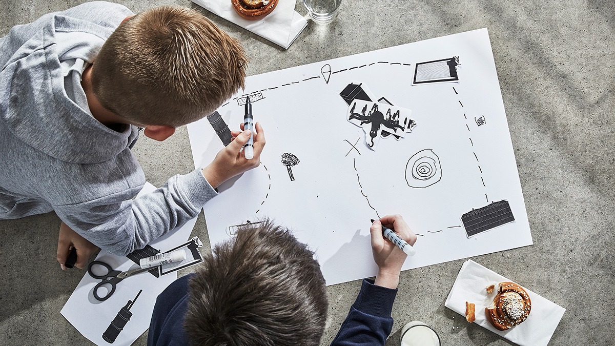 Two children lying on the floor drawing a map. Next to them are cinnamon rolls and crafting tools.
