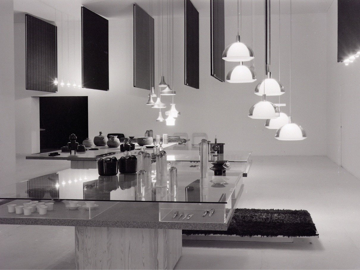 Utställning med stilren design, stort bord i glas och trä, från taket hänger lampor i krom.