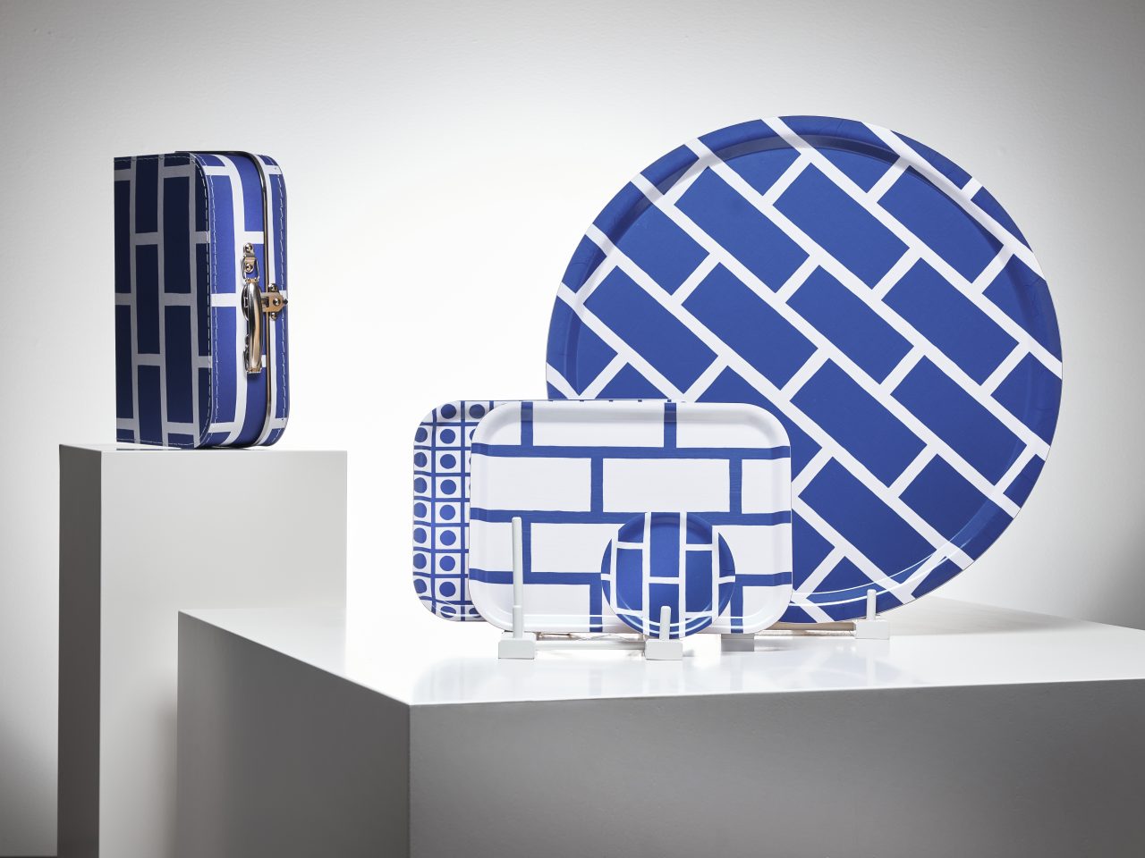 Ett glasunderlägg och brickor i olika storlekar, grafiska mönster och färger, och en liten resväska i blåvitt.