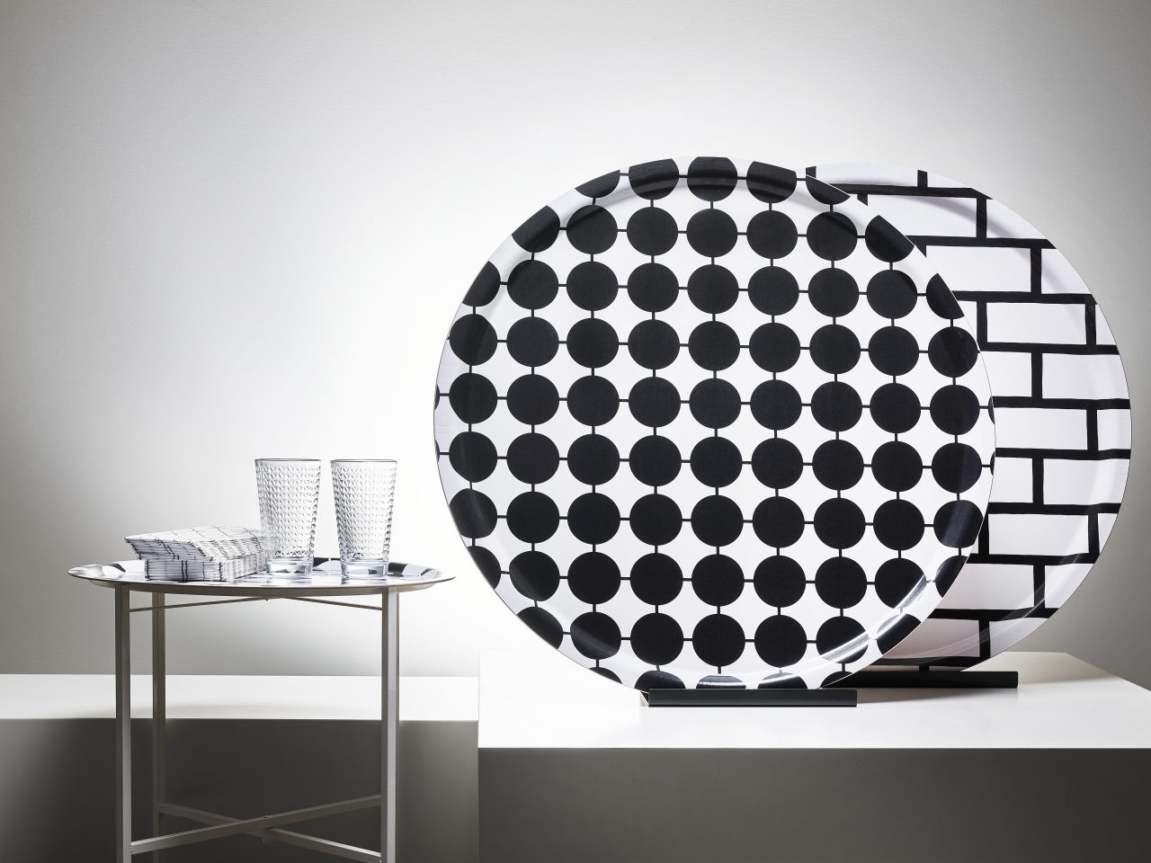 Two glasses, a stack of paper napkins on a small tray table next to two large round trays with black/white graphic patterns.