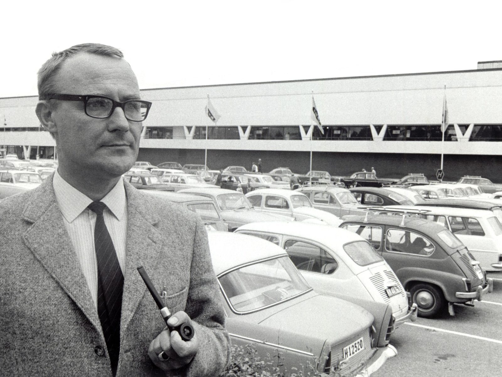 Ingvar Kamprad in Tweedjacke steht vor einem weißen Gebäude, dem ersten IKEA-Einrichtungshaus und einem Parkplatz mit Autos aus den 1950er-Jahren.