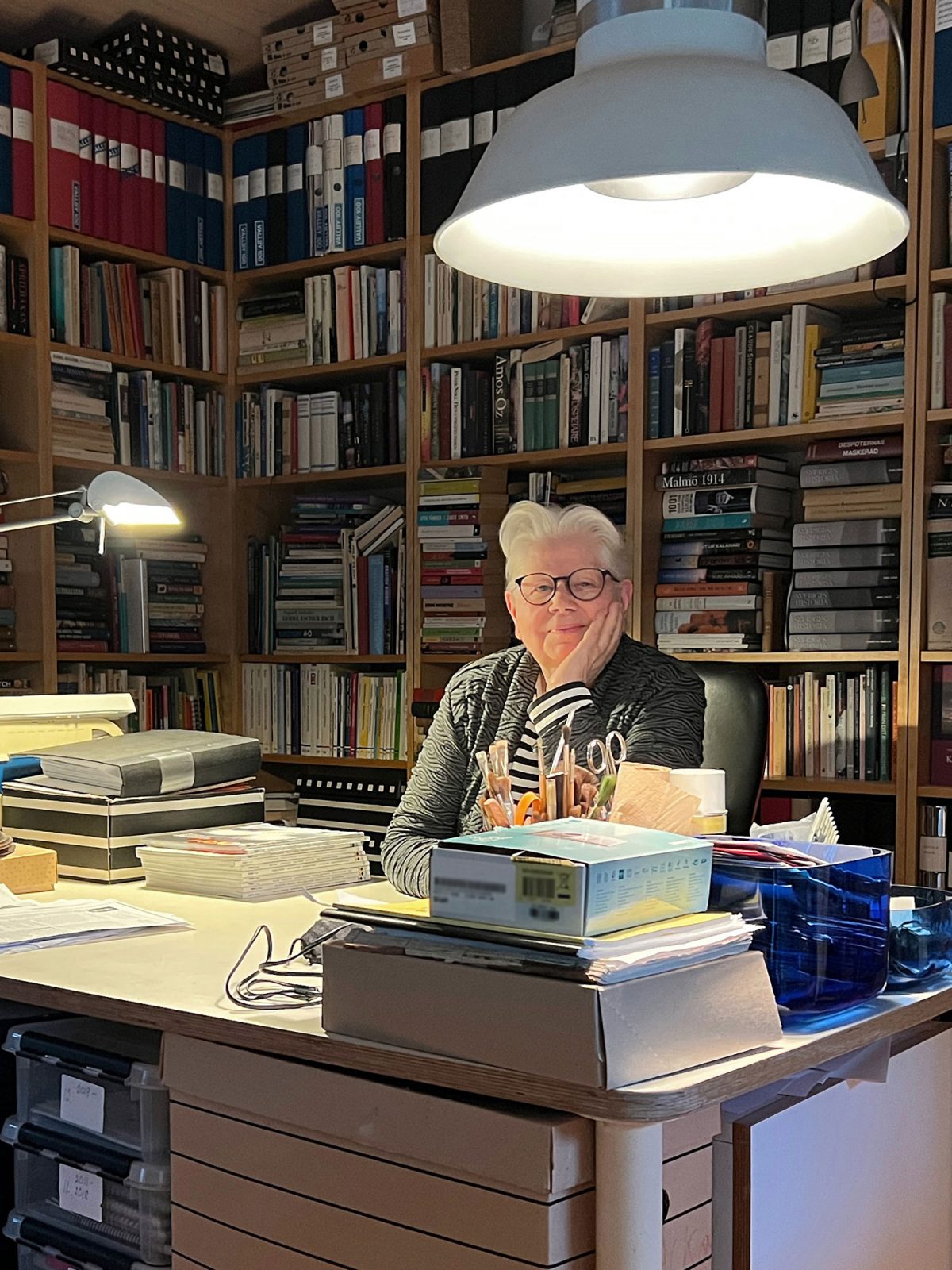Äldre vithårig kvinna i randig tröja, Vivianne Sjölin, sitter bakom skrivbord i överbelamrat arbetsrum.