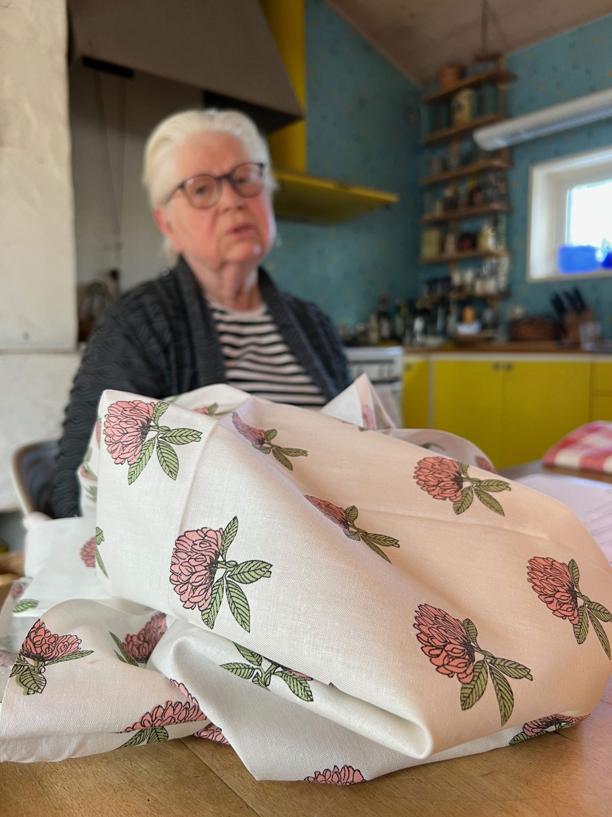 Äldre vithårig kvinna i randig tröja, Vivianne Sjölin, visar en bit tyg med rosa klöverblommor på vit botten.