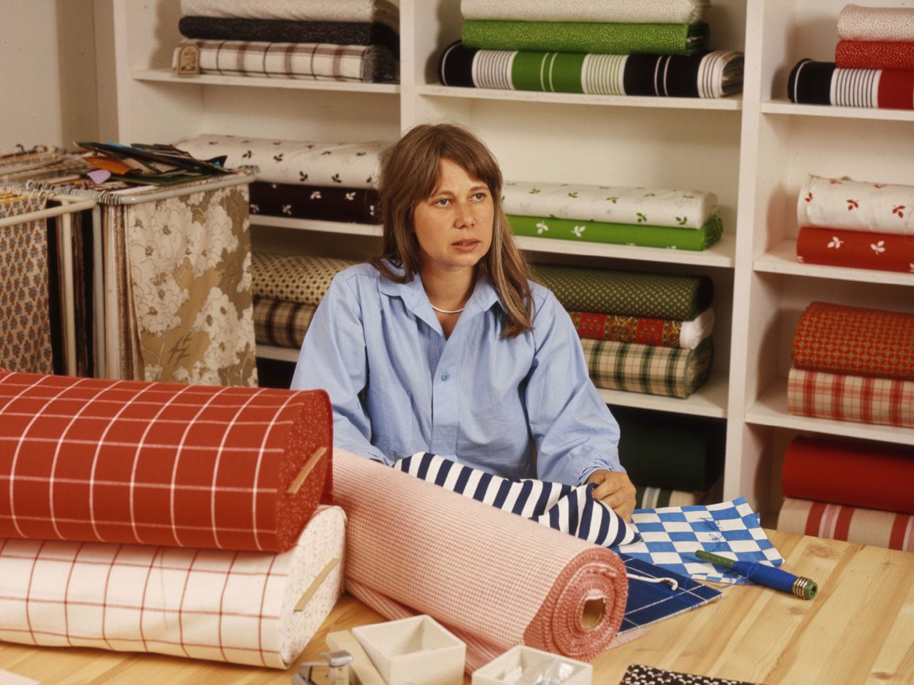 Young woman in blue shirt, Vivianne Sjölin, surrounded by colourful, patterned fabric rolls.