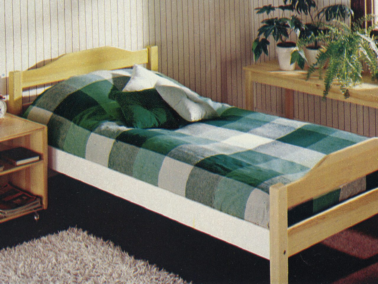 1977 IKEA catalogue image of a wooden bed with green check INDUS bedspread and a white rug on the floor.
