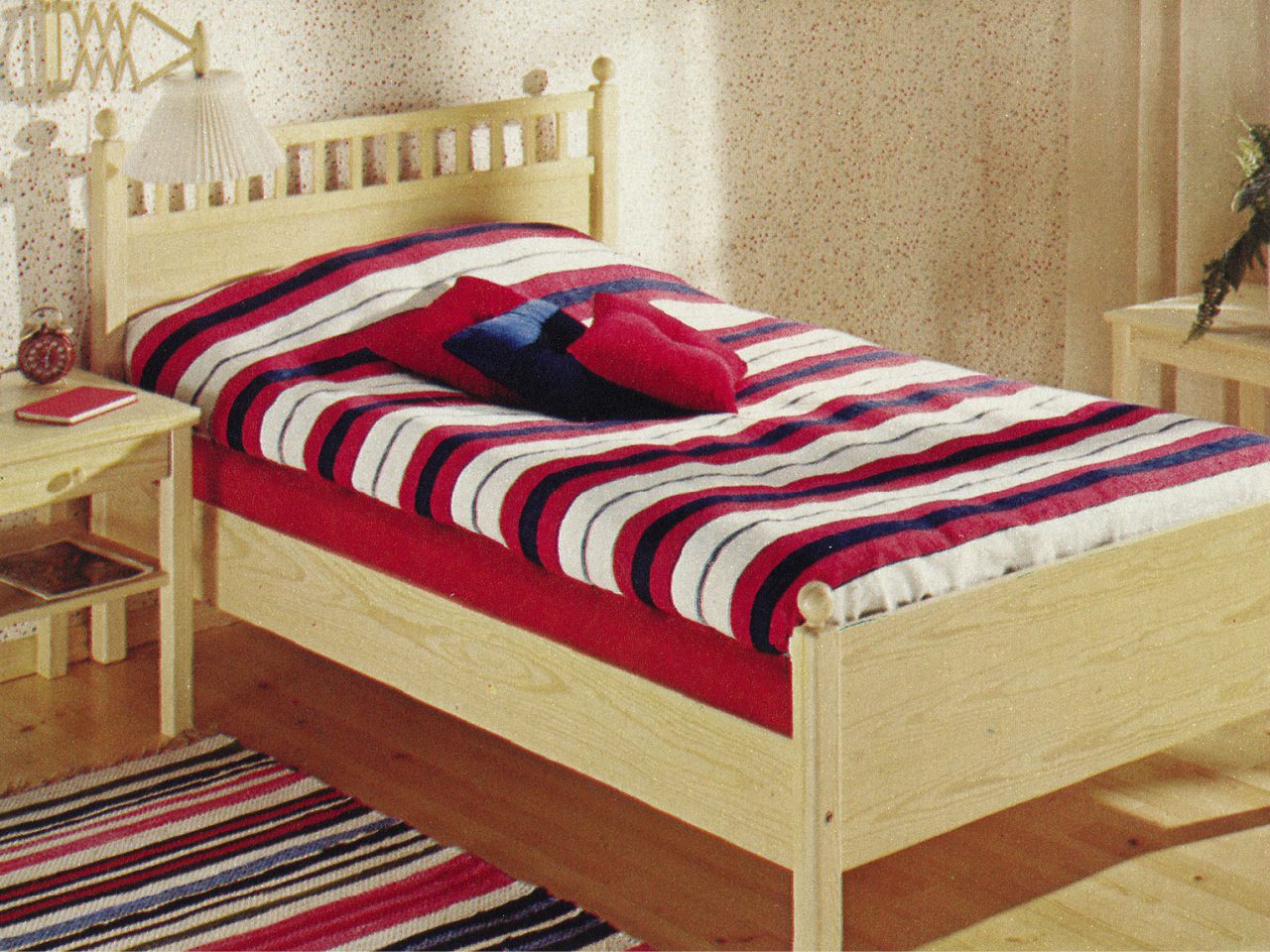 1977 IKEA catalogue image of a wooden bed with a red, black, and white bedspread and a striped rug on the floor.