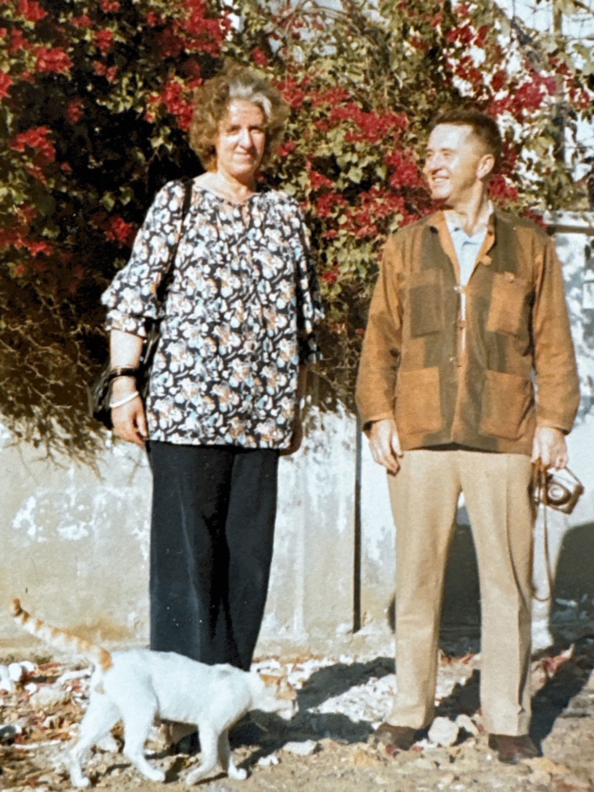Inez Svensson in a patterned blouse with a happy man in a brown jacket; a white cat walks by their feet.
