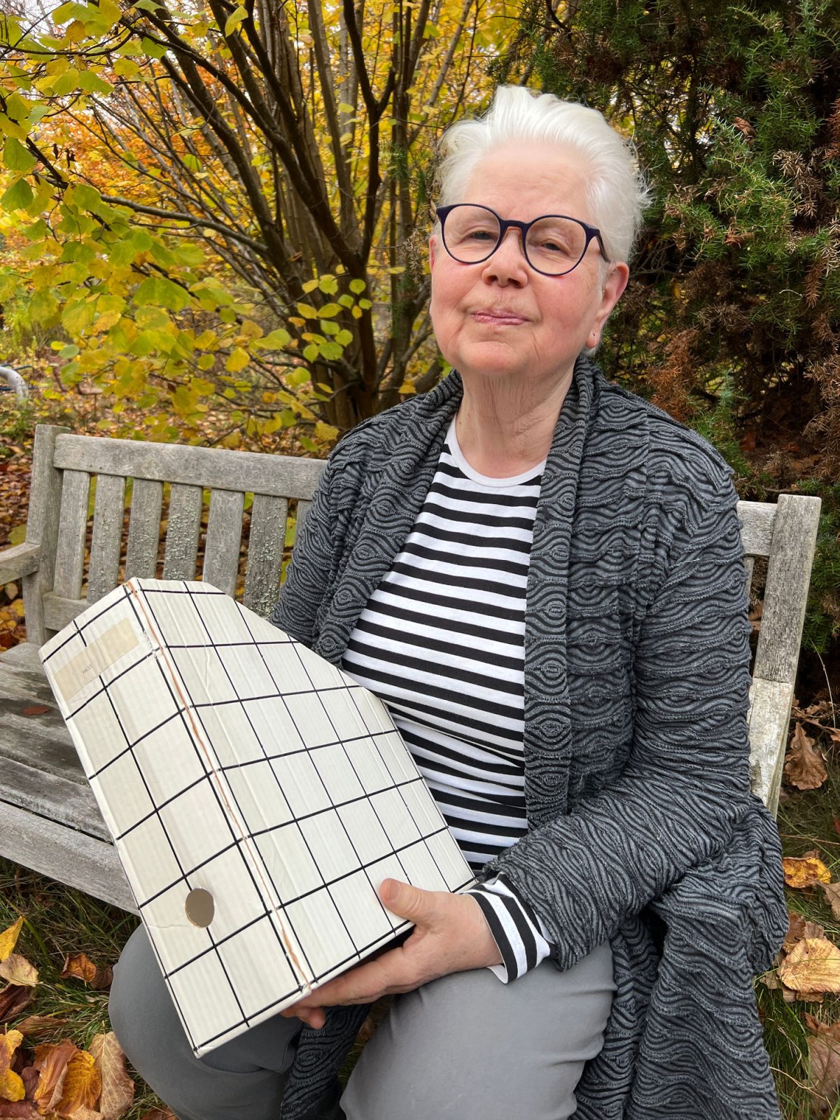 Vithårig kvinna i grå kofta och randig tröja sitter på bänk och håller i tidskriftssamlare med svart-vit-rutigt mönster.