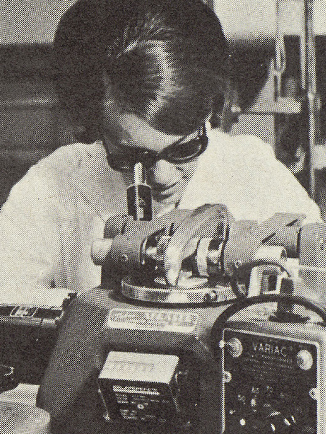 Woman in 1960s-style clothing and hairdo looks into a microscope in a lab environment.