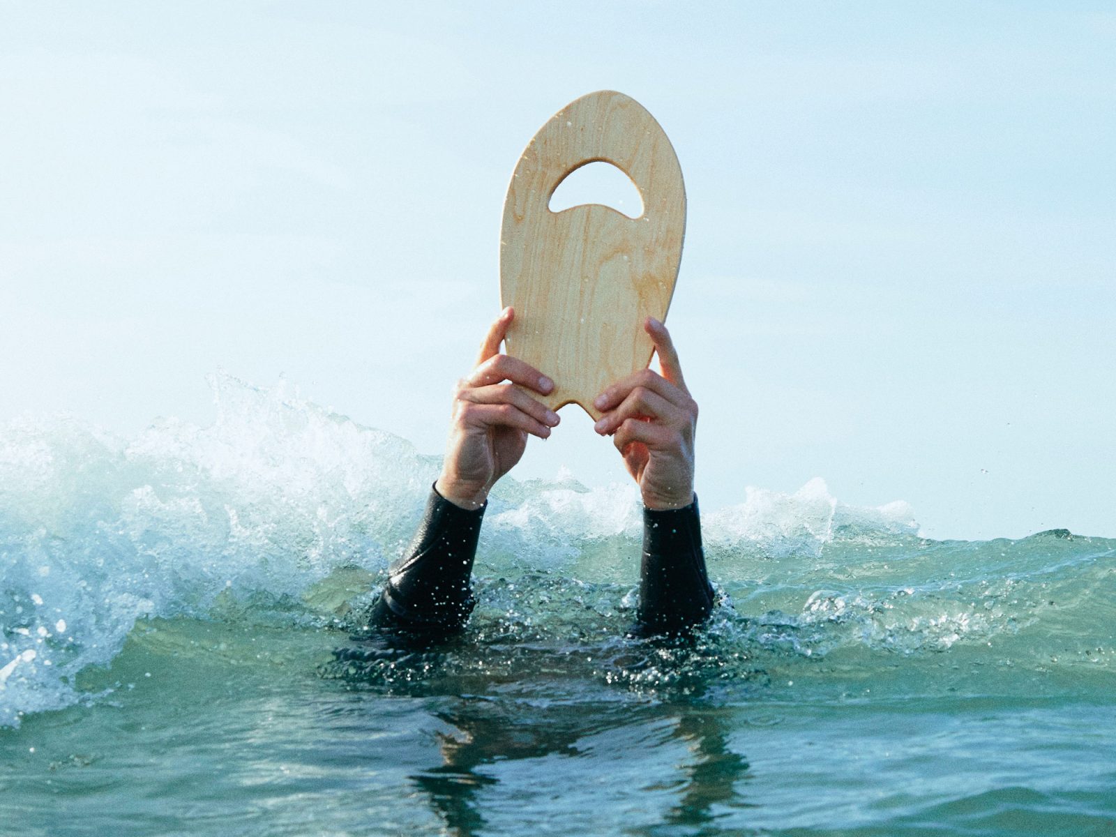 Arms covered by wet suit hold small blond wooden board above water, from IKEA KÅSEBERGA surf collection.
