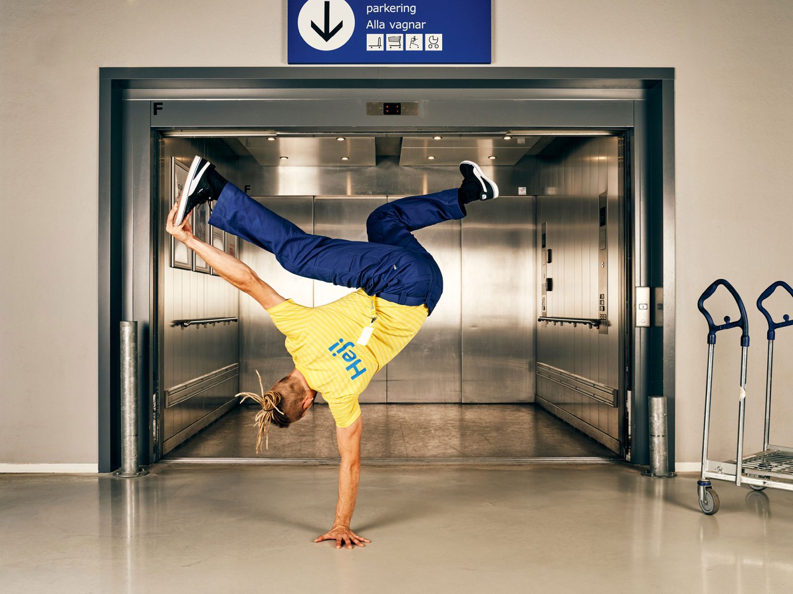 Young blond man in IKEA blue and yellow work clothes does a break dance move in front of an IKEA store elevator.