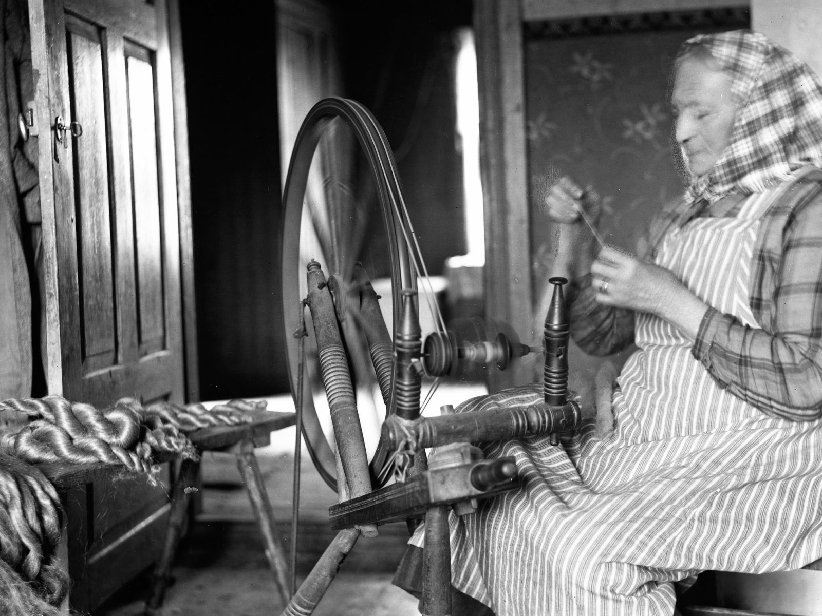 Blonde woman in shawl and striped apron spinning flax on a spinning wheel, 1920s.