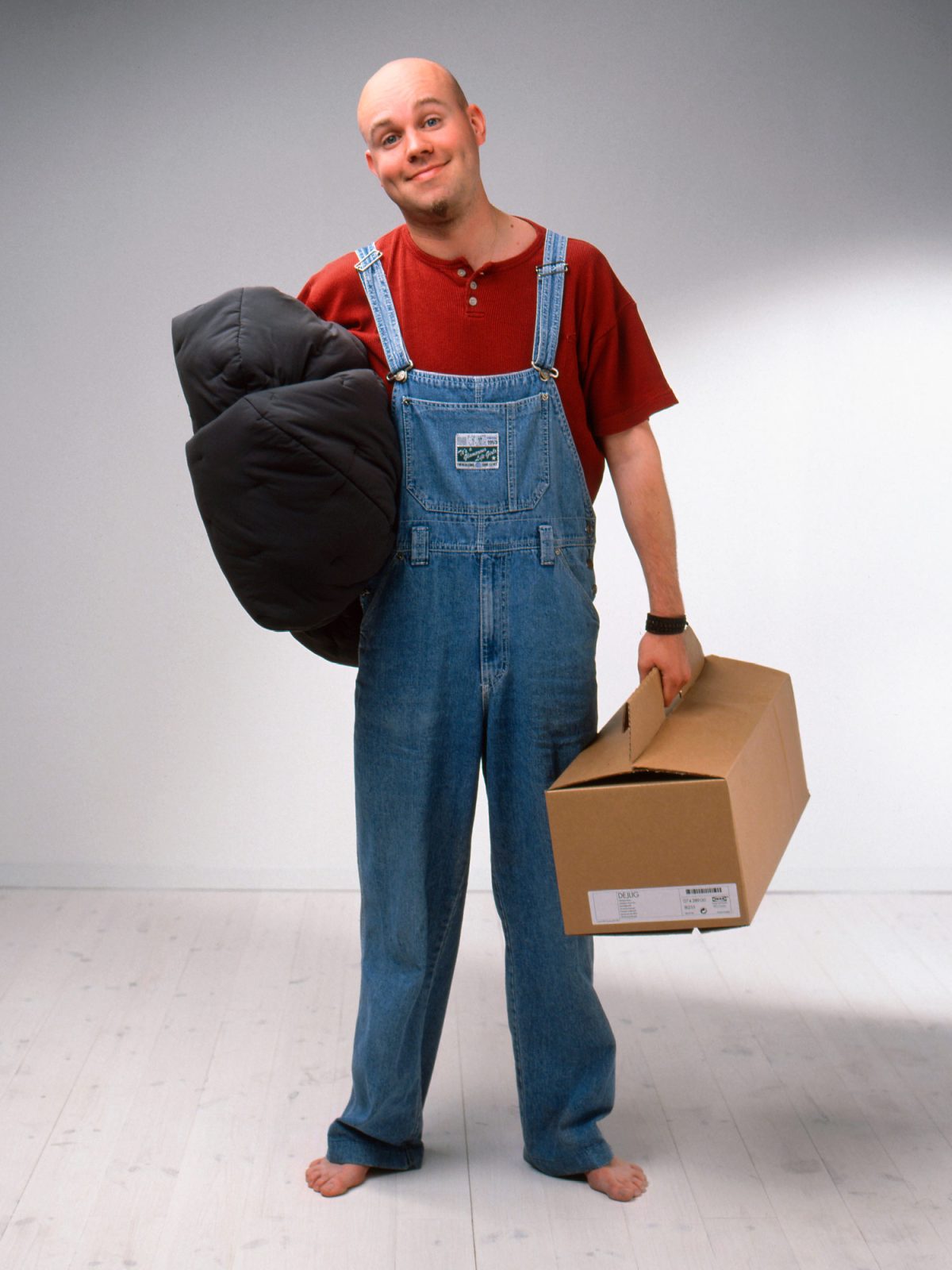 Smiling young man in dungarees holds a bunch of fabric under one arm, in one hand he holds a small brown flat pack.
