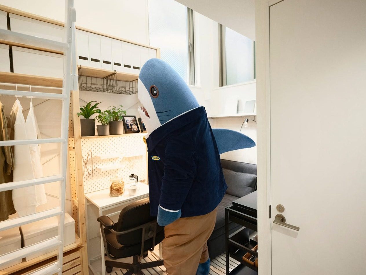 A person dressed in a shark costume and suit standing at a desk under a loft bed.