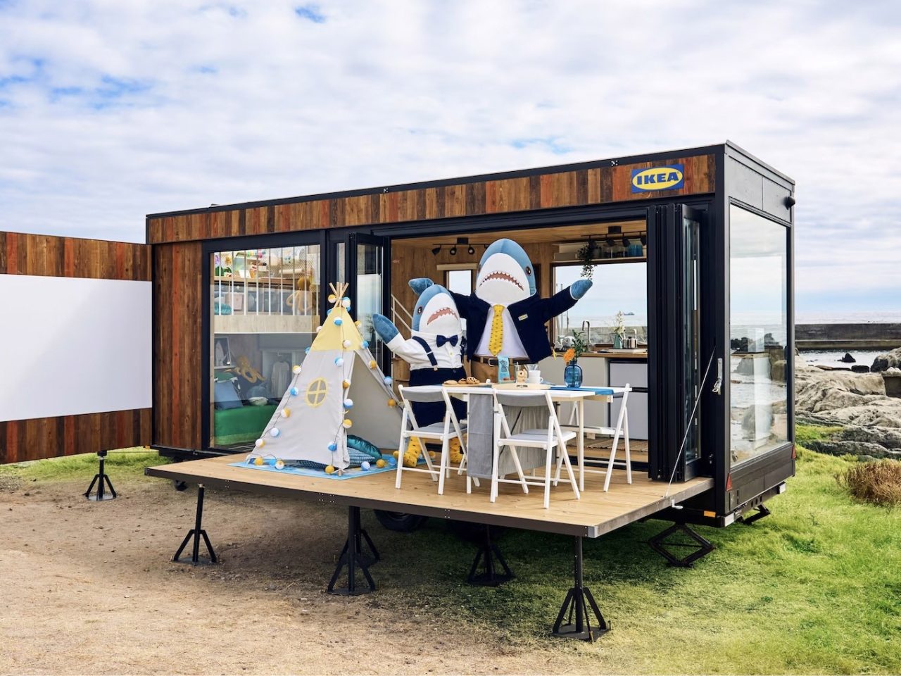 Two people dressed in shark costumes standing in front of a small wooden trailer with an IKEA sign on the wall.