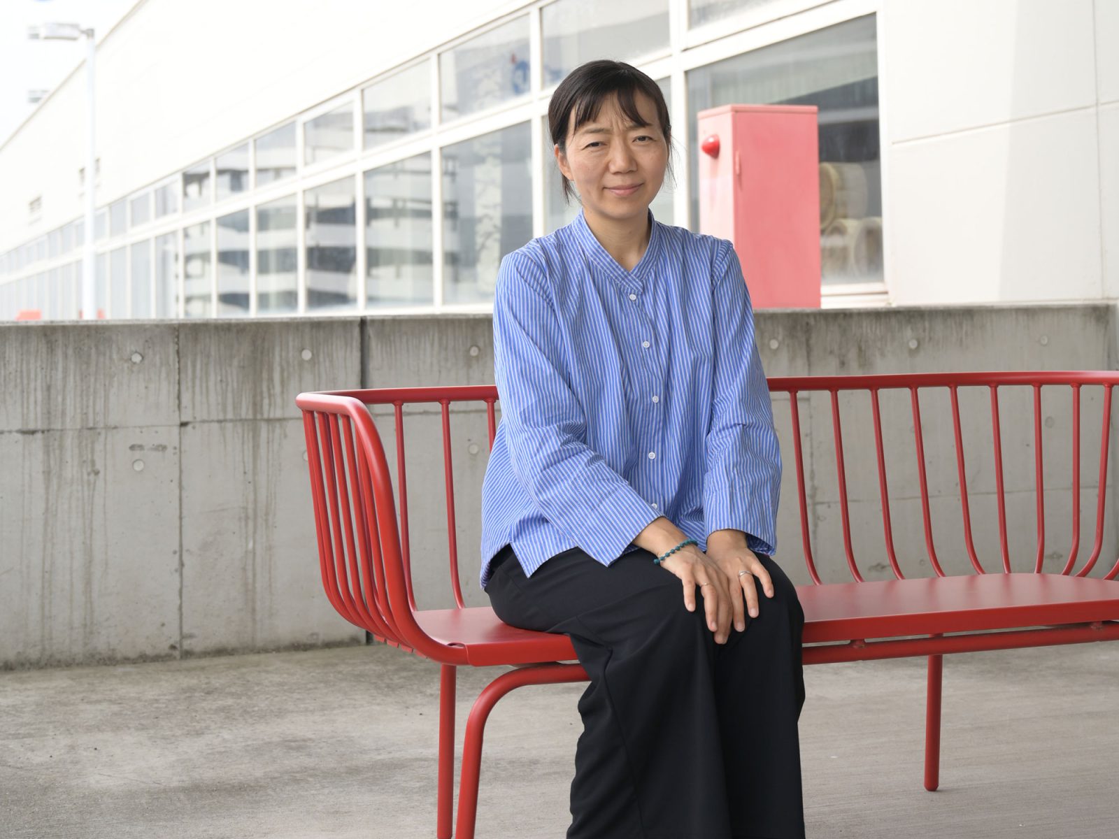 Mörkhårig kvinna i blå blus sitter på en röd metallsoffa, Aya Kosai.