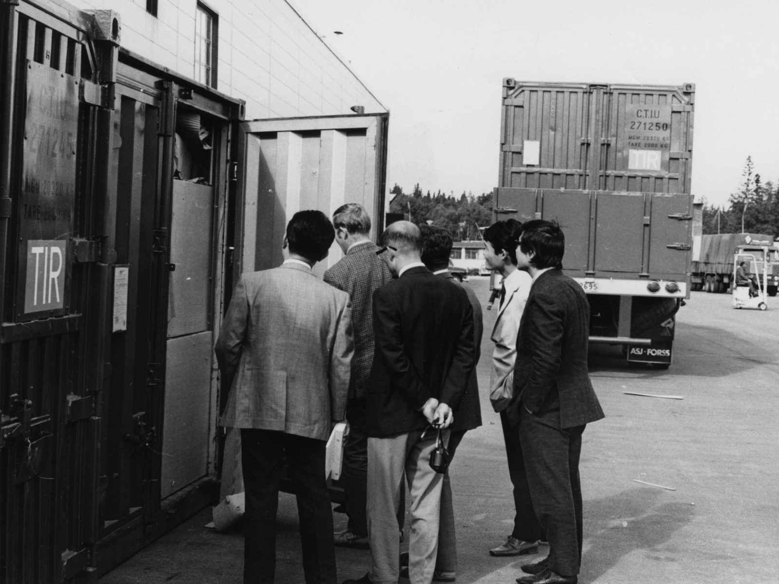A group of men in suits standing in an industrial area, peeking into a container.