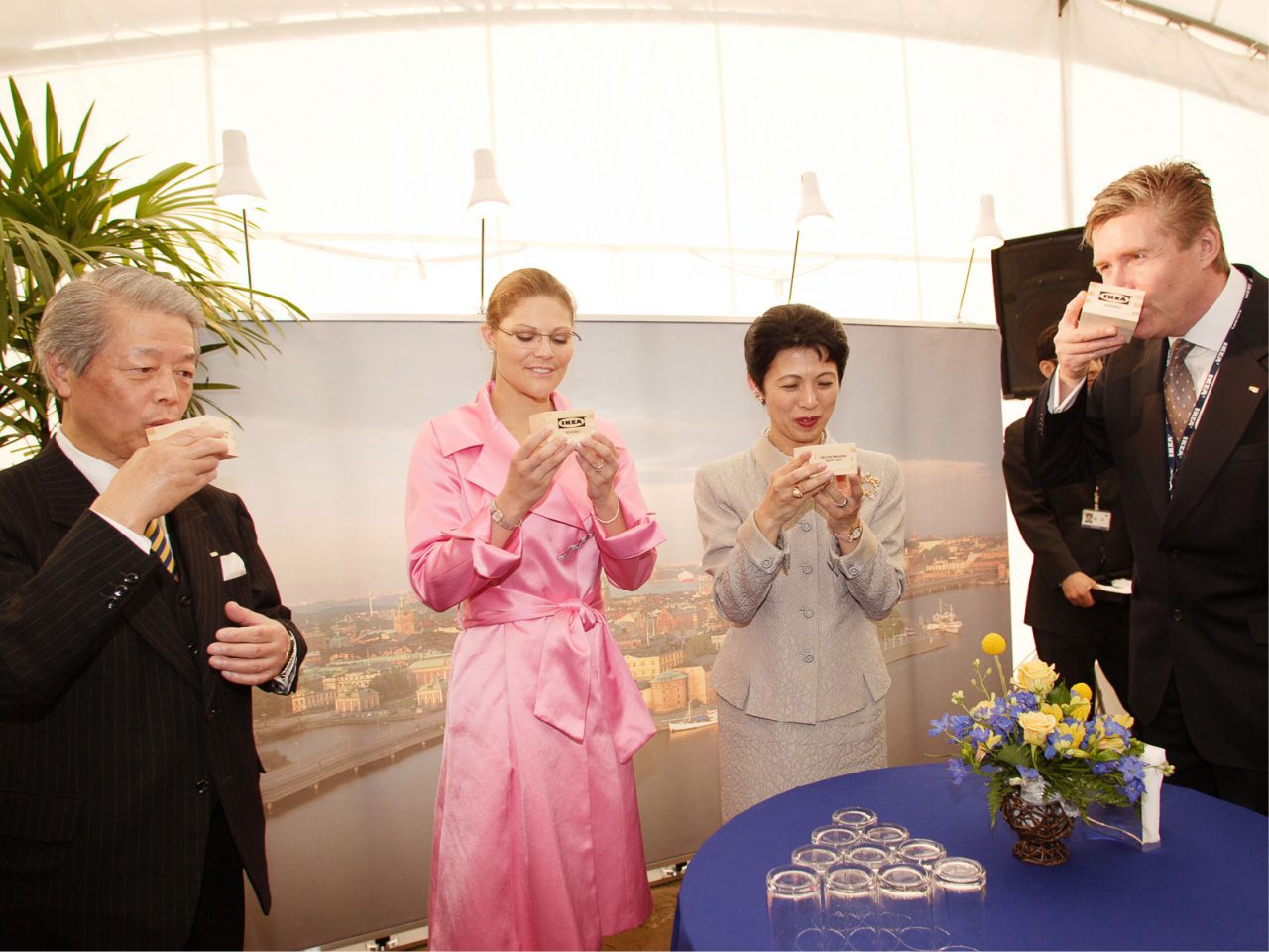 Four well-dressed people, two men in dark suits, woman in pink suit, woman in grey suit drinking from wooden bowls.
