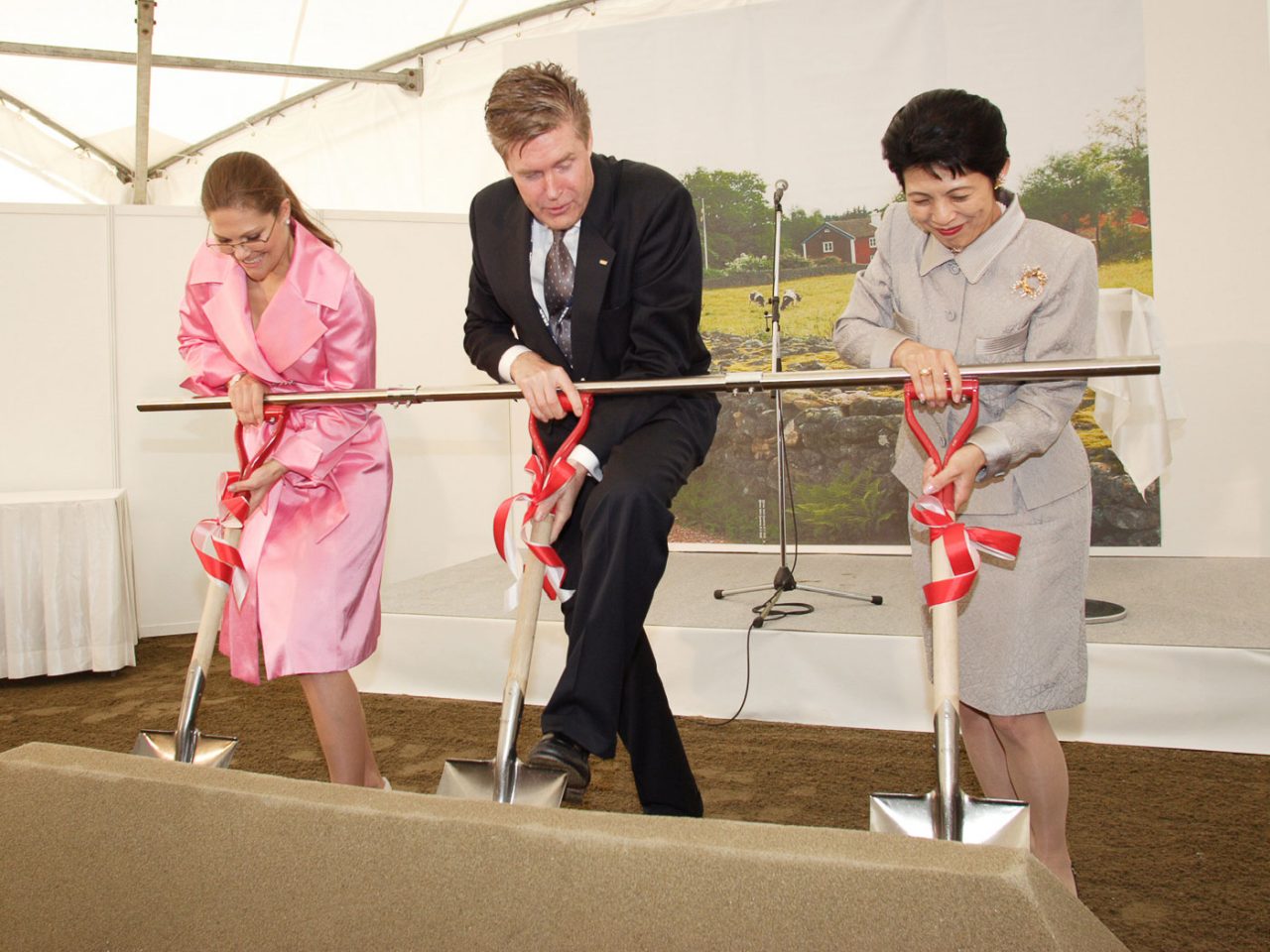 Three well-dressed people, woman in a pink suit, man in a dark suit, woman in a grey suit, each putting shovels into ground.