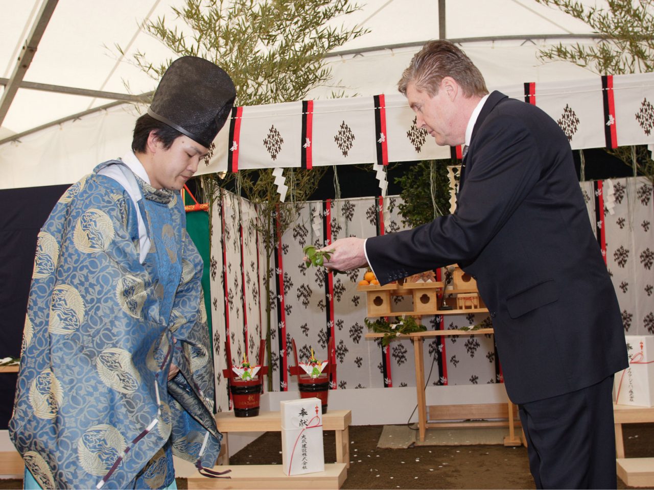 Man in black suit, Tommy Kullberg, gives an olive branch to a Shinto priest in a high hat and blue silk kimono.